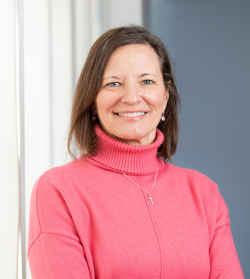 Lori Rolando poses indoors, wearing a bright pink turtleneck sweater, with her arms crossed in front of a window and gray wall.