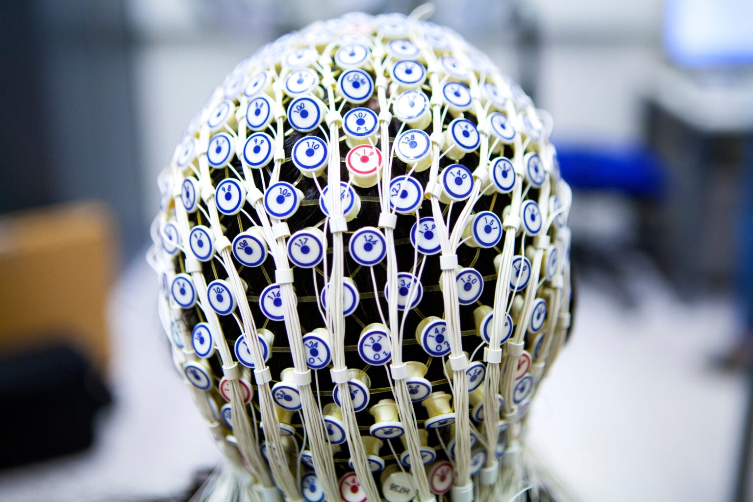 Dome-shaped EEG electrode cap covered with numerous numbered circular sensors and bundled white wires, shown against a blurred laboratory background.