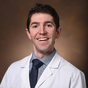 Portrait of a smiling man wearing a white lab coat, patterned shirt, and dark tie against a brown background.