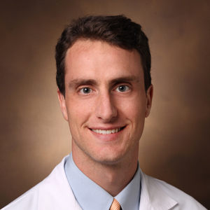 Head-and-shoulders portrait of a smiling man wearing a white medical coat, light blue shirt, and striped tie.