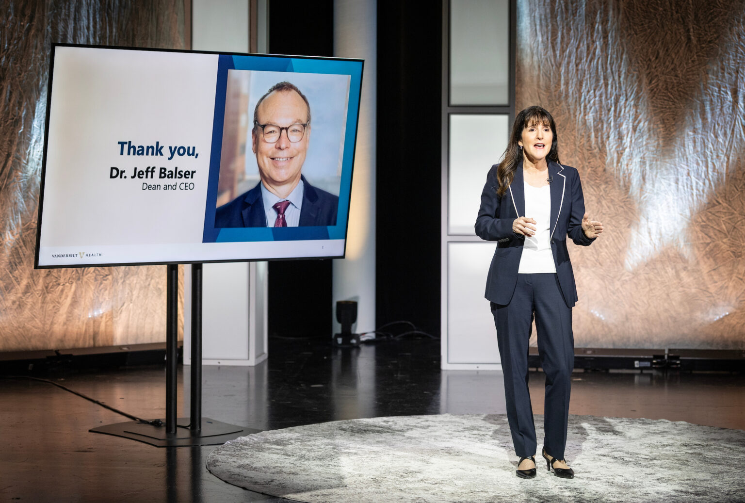 Jane Freedman stands on a studio set, speaking beside a display that reads “Thank you, Dr. Jeff Balser, Dean and CEO” and shows his portrait; Vanderbilt Health branding appears at the bottom.