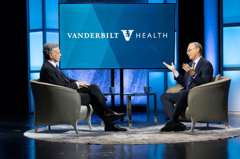Dr. Balser and John Seigenthaler sit in armchairs facing each other on a stage, with a small table between them and a screen displaying the Vanderbilt Health logo behind them.