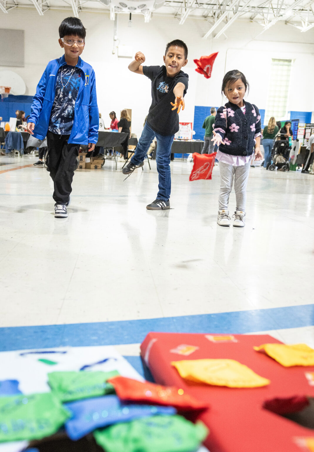 Jose Lajom, Emilio Lajom, and Emily Lajom play a game in a school gym during the MEGAMicrobe event, with other activity tables and colorful game pieces in the foreground.