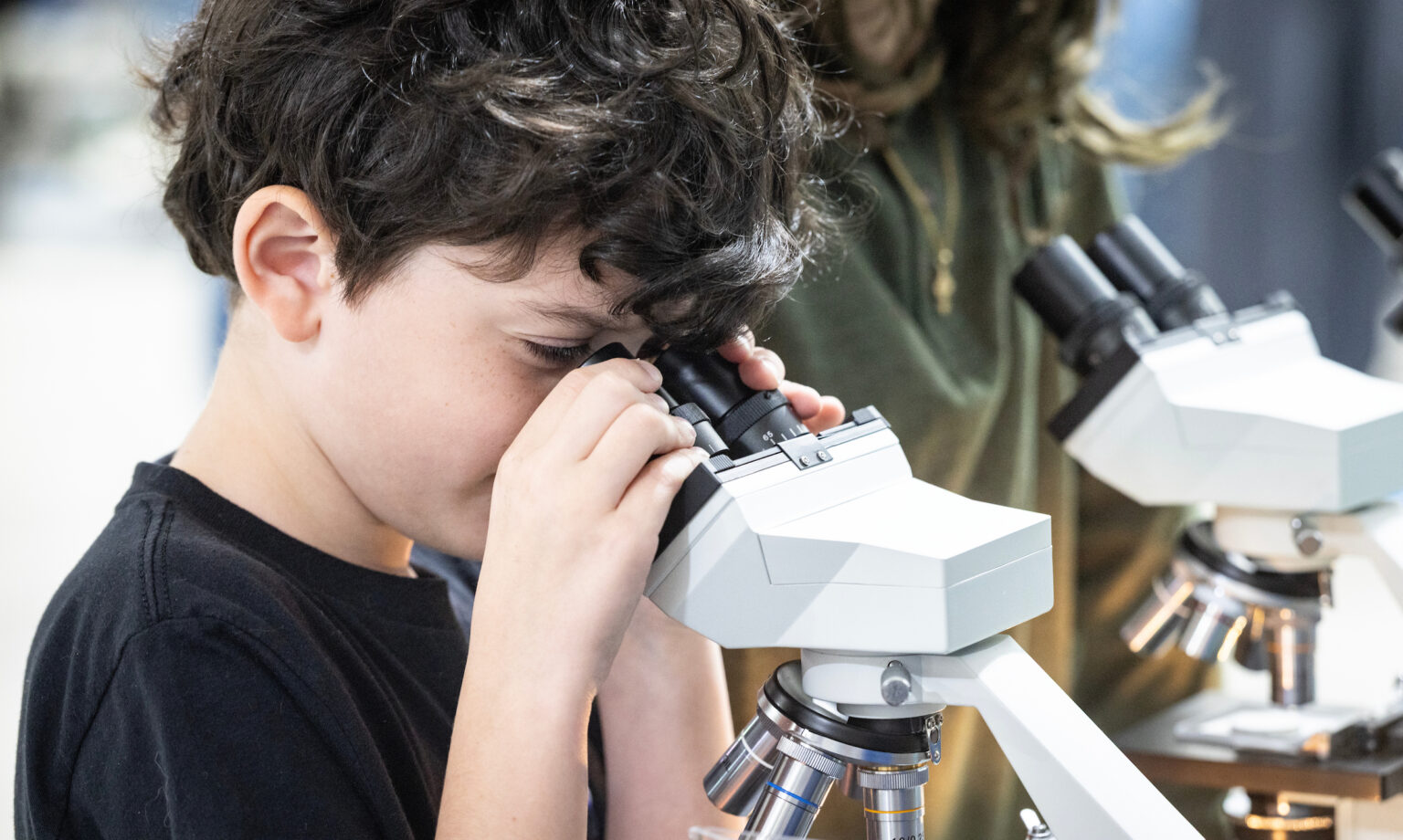Lucas Nelson, a child with dark curly hair, looks through the eyepiece of a white compound microscope while another microscope sits behind him.