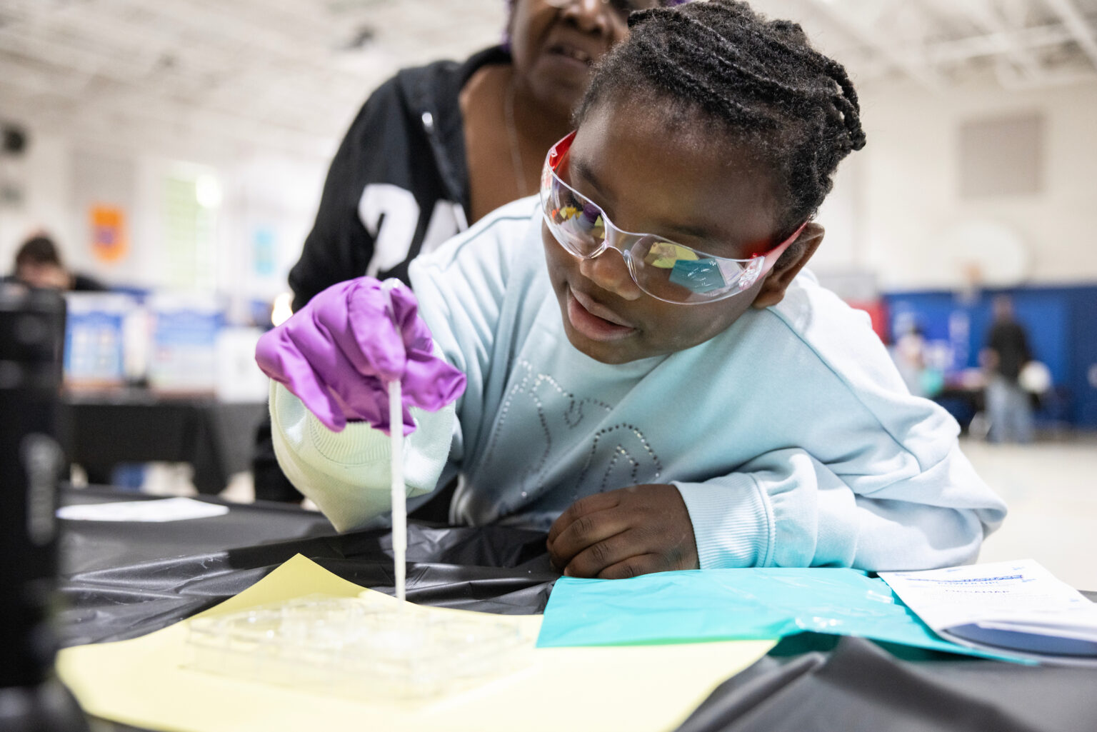 Child wearing safety goggles and a purple glove uses a pipette at a hands-on science demonstration while an adult assists from behind.