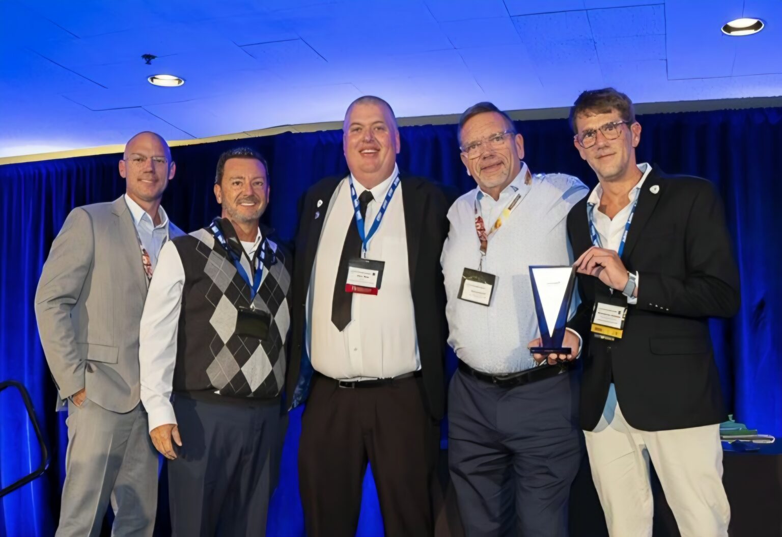 Five men wearing conference badges pose together in front of blue curtains; the man second from the right holds a triangular glass award.