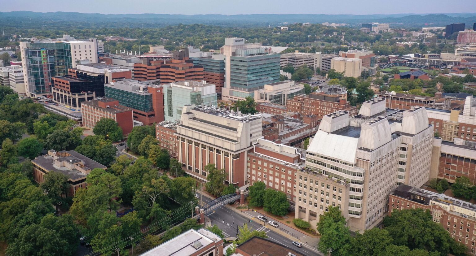 Aerial view of Vanderbilt University Medical Center and surrounding Vanderbilt University campus buildings in Nashville, with wooded hills and city neighborhoods in the background.