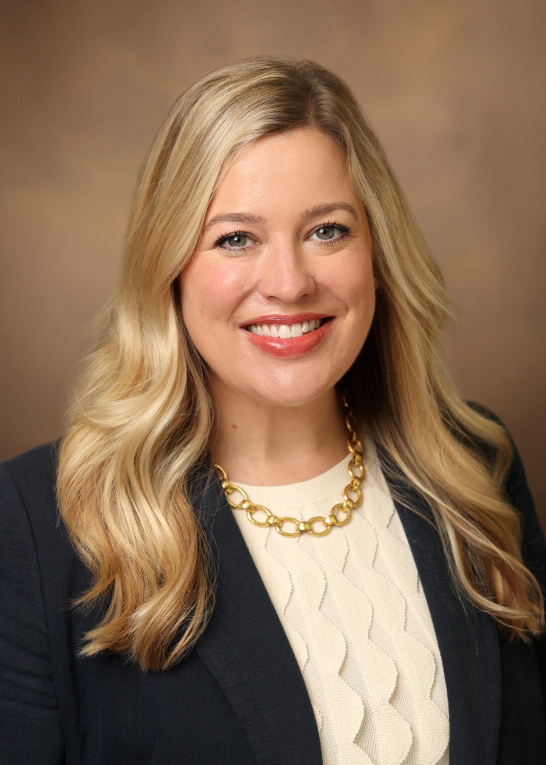 Portrait of Rebecca Warren smiling at the camera, with long wavy blonde hair, wearing a black blazer, cream textured top, and gold chain necklace against a warm brown background.