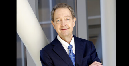John Howser poses with folded arms in a navy suit and blue patterned tie in a modern building interior.