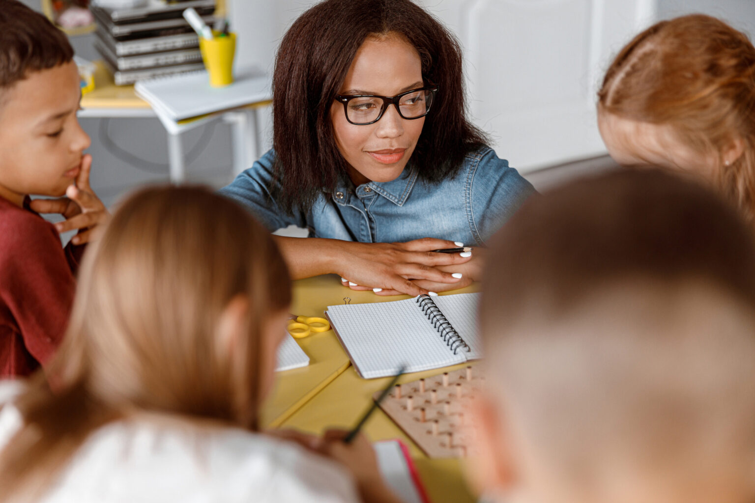 A teacher wearing glasses and a denim shirt sits at a classroom table with several children, who are gathered around notebooks and school supplies.