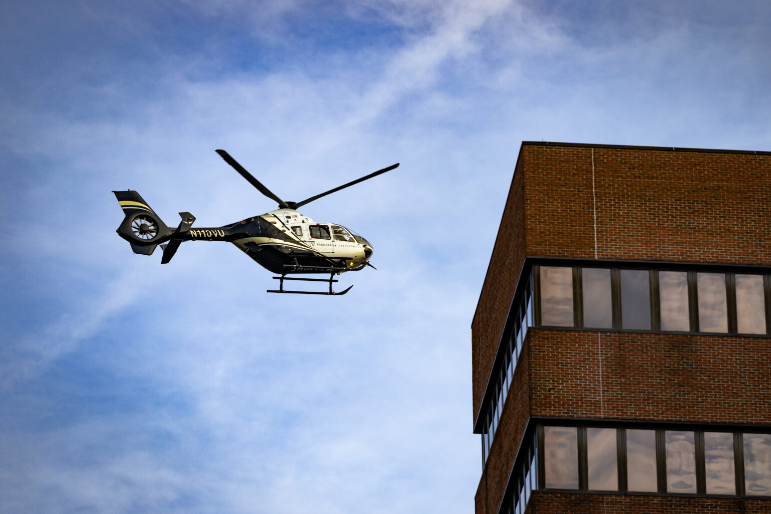 A black-and-white medical helicopter marked “Vanderbilt LifeFlight” flies beside a brick hospital building under a partly cloudy blue sky.