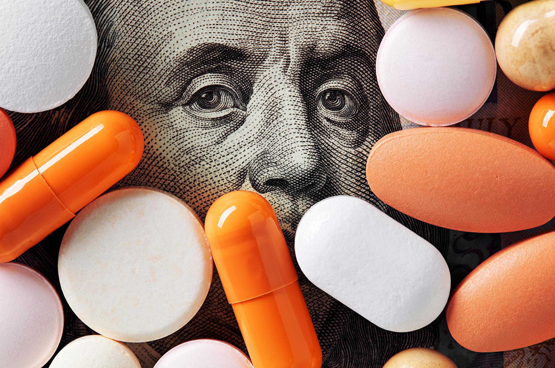 Close-up of assorted orange, white, pink, and peach pills and capsules arranged over a U.S. $100 bill.